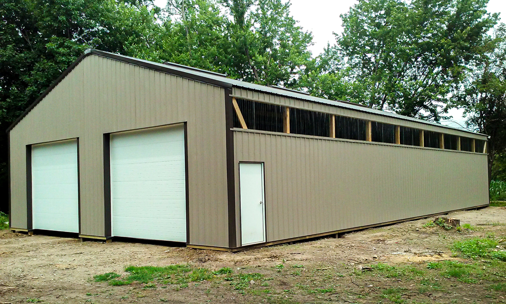 A large, rectangular metal building with two closed garage doors and a single entrance door. The structure has small windows just below the roof and is surrounded by dirt and trees in the background.