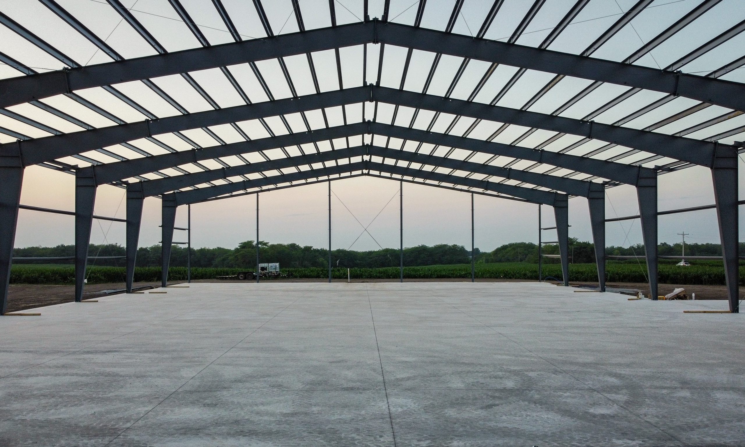 Partially constructed large metal building frame with steel beams, standing on a concrete foundation. The surrounding area is outdoors with green fields and trees visible in the background. The sky is clear with early morning or late afternoon light