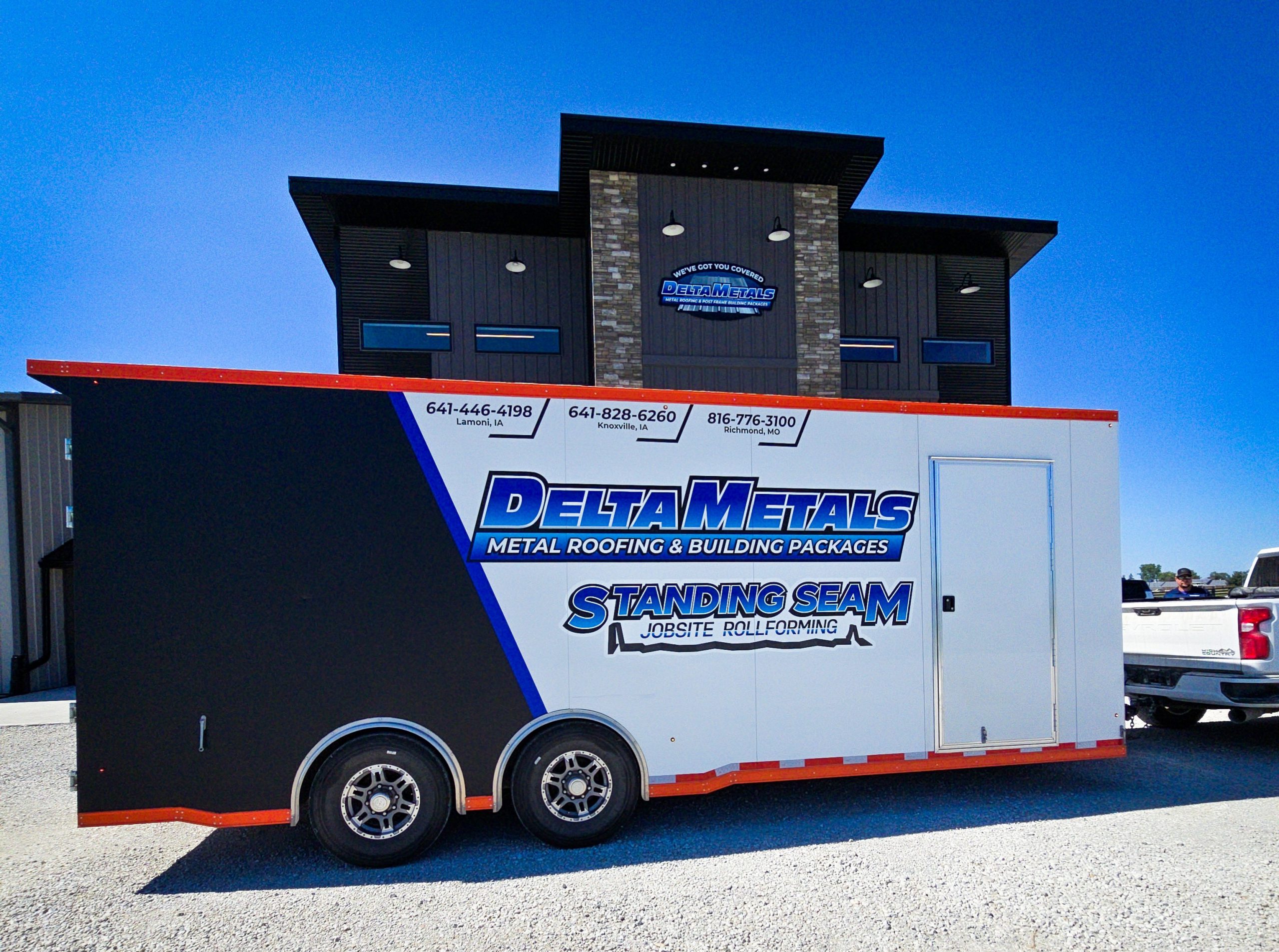 A large white trailer with black and orange accents features the logo and contact information for Delta Metals, a metal roofing and building packages company. The trailer is parked in front of a modern commercial building under a clear blue sky.