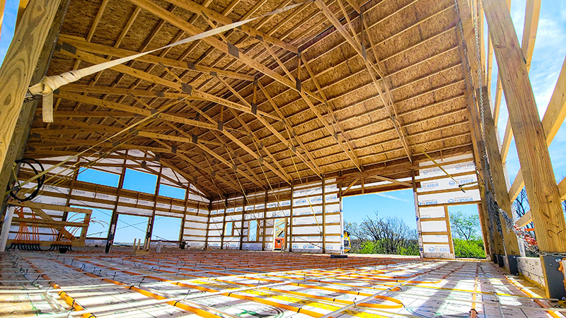 Interior view of a partially constructed building with a wooden frame and high ceiling. The floor shows pipes and wiring, indicating ongoing work. Sunlight streams in through the open walls, highlighting the structure's framework.