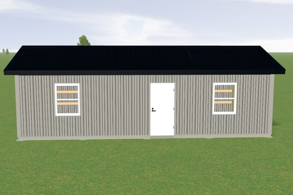 A small, gray building with a black roof stands on a green field. The structure features a white door and two windows with four horizontal panes each. The sky is mostly clear with a few distant trees visible.