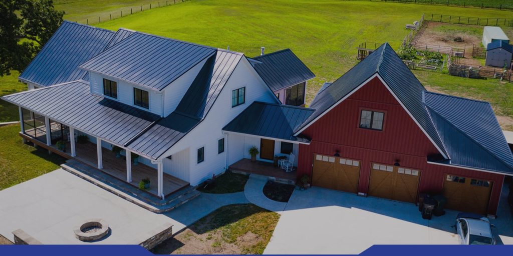 Aerial view of a modern farmhouse with a white main section, a red barn-style garage, metal roof, wide porch, and a large green lawn with a garden and outbuildings in the background.