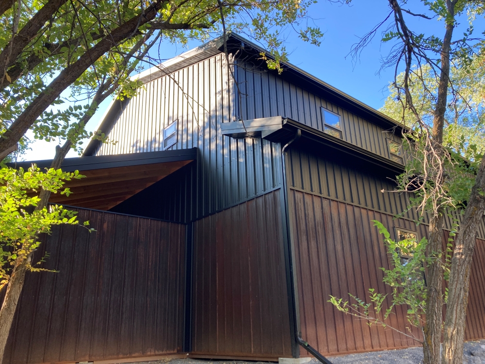 A modern dark brown house with metal siding and a steep roof stands among leafy green trees under a clear blue sky.